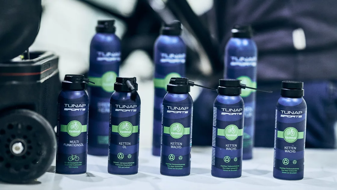 Mehrere dunkelblaue Spraydosen von TUNAP SPORTS für Fahrräder stehen auf einer hellen Fläche. Die Dosen sind mit einem grünen Etikett versehen, auf dem ein Fahrrad abgebildet ist und die jeweilige Funktion des Produkts angegeben ist: Multifunktionsöl, Kettenöl und Kettenwachs. Die Vorderseite der Dosen zeigt den Markennamen TUNAP SPORTS und das Wort „FAHRRAD“. Die Produktbeschreibungen betonen Eigenschaften wie Langlebigkeit, Schmutz- und Wasserabweisung, sowie Schutz vor Verschleiß. Der Hintergrund ist unscharf und zeigt einen Teil einer dunklen Tasche oder eines Beutels. Das Bild vermittelt einen Eindruck von Sauberkeit, Funktionalität und hochwertigen Fahrradpflegeprodukten. © Sport Kessler GmbH