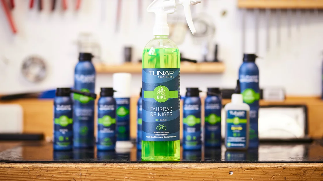 Eine Sprühflasche mit Fahrradreiniger von TUNAP SPORTS steht im Vordergrund des Bildes. Die Flüssigkeit in der Flasche ist grün. Der Hintergrund zeigt eine unscharfe Werkstatt mit Werkzeugen an der Wand. Mehrere weitere Flaschen mit Fahrradpflegeprodukten von TUNAP SPORTS sind im Hintergrund unscharf zu sehen, was auf eine Produktreihe hindeutet. Das Bild vermittelt Sauberkeit, Funktionalität und eine professionelle Arbeitsumgebung. Der Fokus auf die einzelne Sprühflasche hebt das Produkt hervor und suggeriert Effizienz und einfache Anwendung bei der Fahrradreinigung. Die Farbgebung in Grün und Blau wirkt beruhigend und professionell. © TUNAP chemisch-technische Produkte Produktions- und Handelsgesellschaft m.b.H.