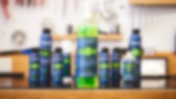 Eine Sprühflasche mit Fahrradreiniger von TUNAP SPORTS steht im Vordergrund des Bildes. Die Flüssigkeit in der Flasche ist grün. Der Hintergrund zeigt eine unscharfe Werkstatt mit Werkzeugen an der Wand. Mehrere weitere Flaschen mit Fahrradpflegeprodukten von TUNAP SPORTS sind im Hintergrund unscharf zu sehen, was auf eine Produktreihe hindeutet. Das Bild vermittelt Sauberkeit, Funktionalität und eine professionelle Arbeitsumgebung. Der Fokus auf die einzelne Sprühflasche hebt das Produkt hervor und suggeriert Effizienz und einfache Anwendung bei der Fahrradreinigung. Die Farbgebung in Grün und Blau wirkt beruhigend und professionell. © TUNAP chemisch-technische Produkte Produktions- und Handelsgesellschaft m.b.H.