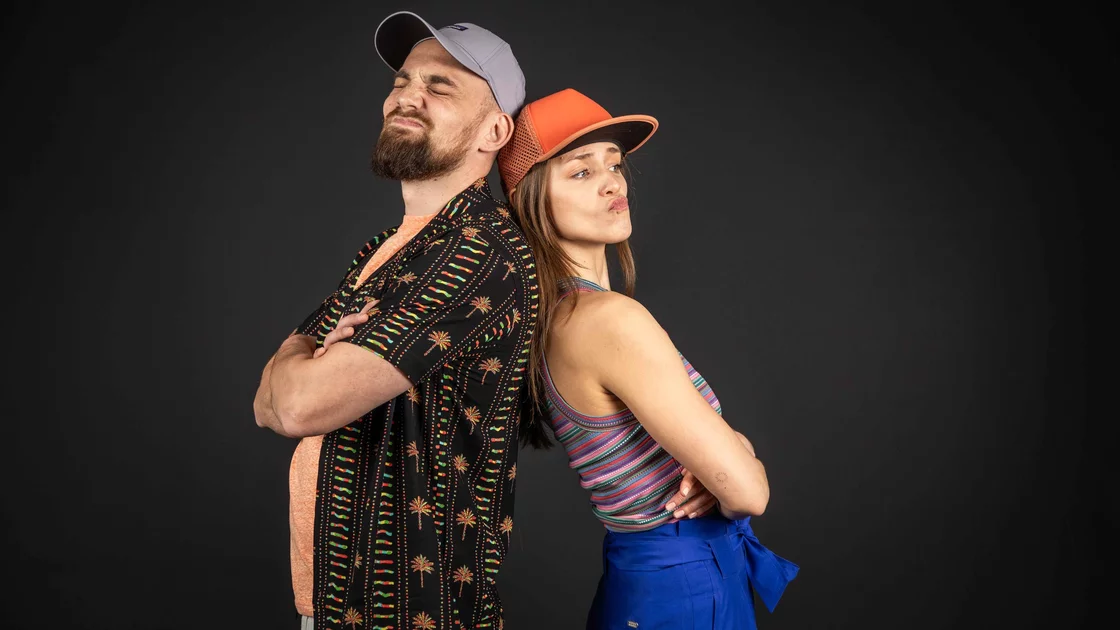 A man and woman stand back-to-back, arms crossed, against a dark background. They both wear baseball caps. © Sport Kessler GmbH