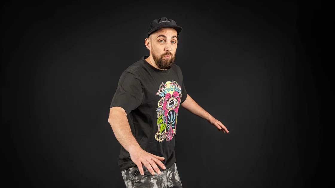 Portrait of a bearded man in a black t-shirt and patterned pants against a dark background. © Sport Kessler GmbH