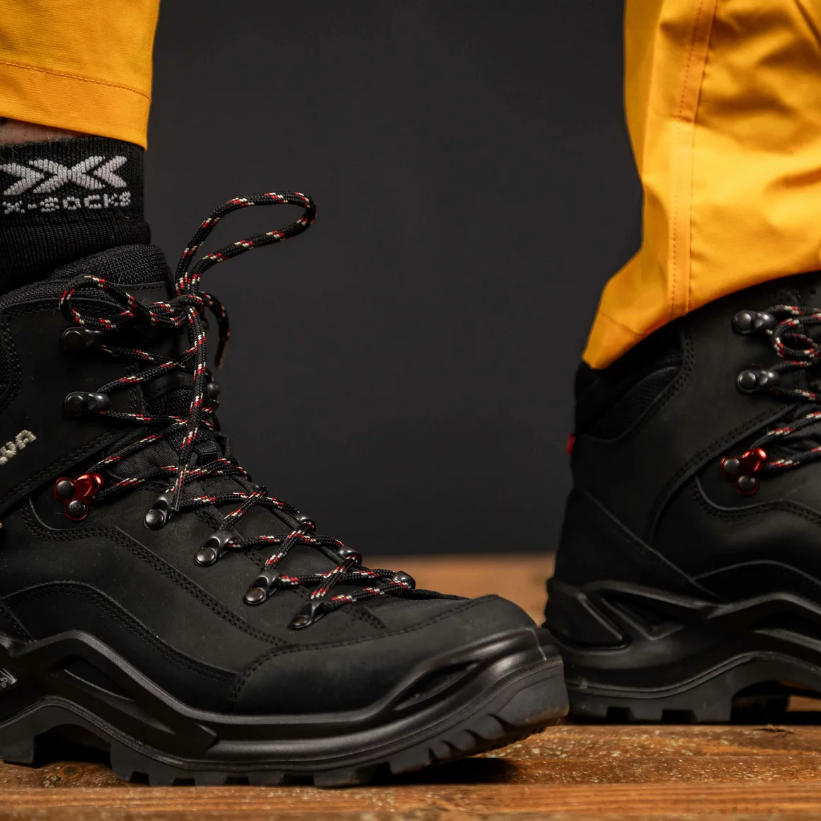 Schwarze Wanderschuhe mit roten und weißen Schnürsenkeln auf einem Holzboden. © Sport Kessler GmbH