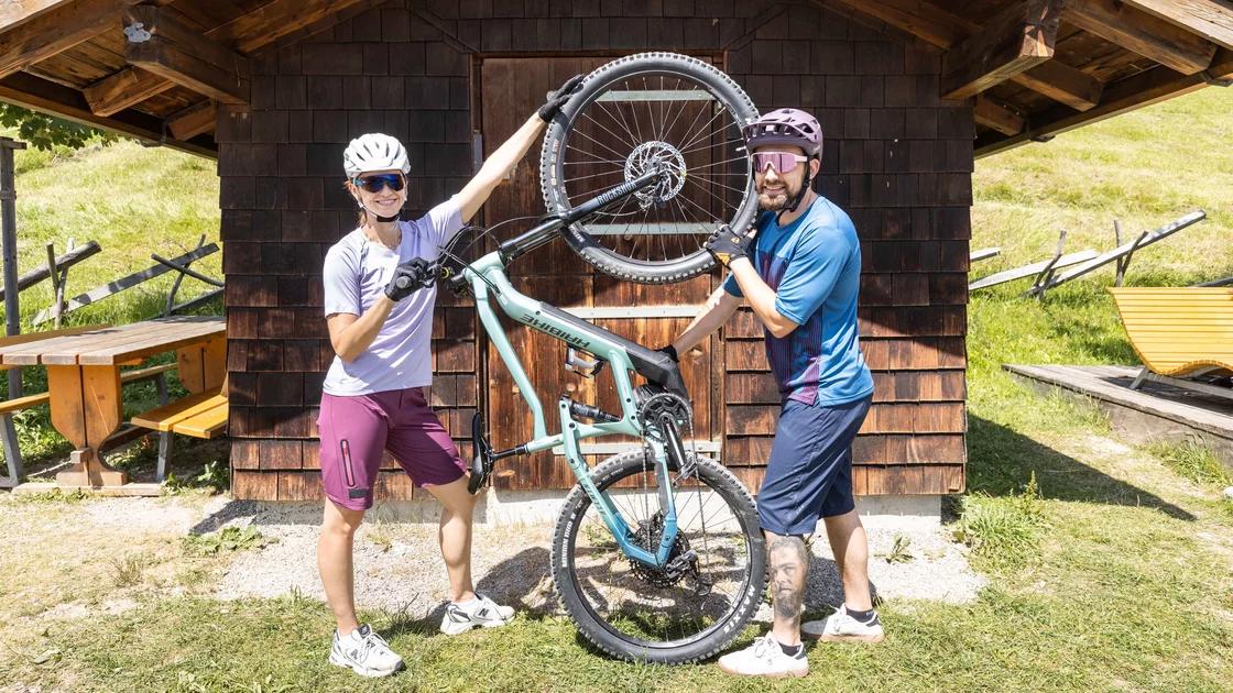Ein Paar hebt ein blaugrünes Mountainbike vor einer Holzhütte hoch. © Sport Kessler GmbH