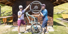 Ein Paar hebt ein blaugrünes Mountainbike vor einer Holzhütte hoch. © Sport Kessler GmbH