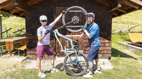 Ein Paar hebt ein blaugrünes Mountainbike vor einer Holzhütte hoch. © Sport Kessler GmbH