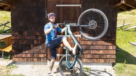 Man with a teal e-mountain bike leaning against a wooden hut. © Sport Kessler GmbH