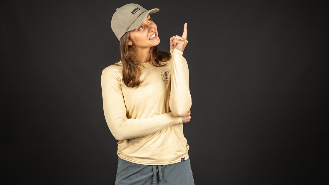 Eine junge Frau mit einer hellbeige Langarm-Oberteil und einem dunkelgrauen Rock vor dunklem Hintergrund. Sie trägt eine hellbeige Baseballkappe und zeigt mit dem Zeigefinger nach oben. © Sport Kessler GmbH