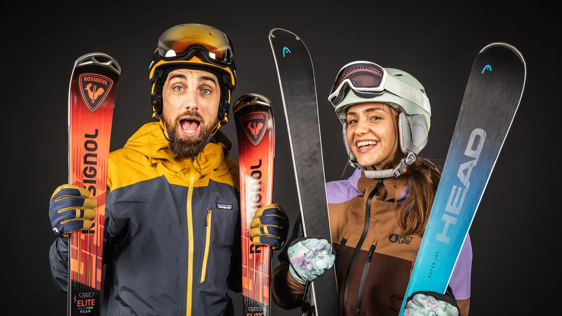 Ein Mann und eine Frau, beide in Skikleidung, posieren vor einem dunklen Hintergrund. Der Mann trägt eine gelbe und dunkelblaue Jacke, eine Skibrille und einen Helm. Er hält einen roten Rossignol-Ski. Die Frau trägt eine braune und lilafarbene Jacke, eine Skibrille und einen Helm. Sie hält einen blauen Head-Ski. Beide lächeln in die Kamera und wirken begeistert und bereit für den Wintersport. Das Bild strahlt Freude und Energie aus und suggeriert die Vorfreude auf einen Tag auf der Piste. © Sport Kessler GmbH