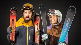 Ein Mann und eine Frau, beide in Skikleidung, posieren vor einem dunklen Hintergrund. Der Mann trägt eine gelbe und dunkelblaue Jacke, eine Skibrille und einen Helm. Er hält einen roten Rossignol-Ski. Die Frau trägt eine braune und lilafarbene Jacke, eine Skibrille und einen Helm. Sie hält einen blauen Head-Ski. Beide lächeln in die Kamera und wirken begeistert und bereit für den Wintersport. Das Bild strahlt Freude und Energie aus und suggeriert die Vorfreude auf einen Tag auf der Piste. © Sport Kessler GmbH