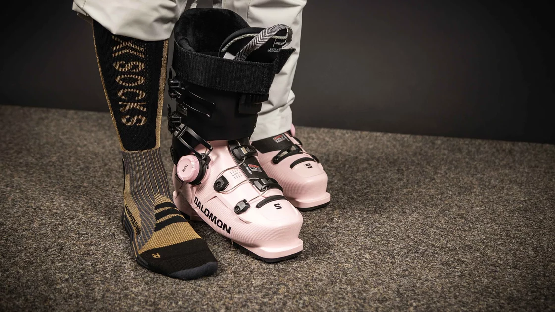 Ein Paar rosafarbene Salomon Skischuhe steht auf einem dunkelbraunen Teppich. Ein Bein, bekleidet mit einer hellgrauen Skihose und schwarzen Socken mit goldfarbener „XSOCKS“-Beschriftung, ist sichtbar. Der Skischuh ist geöffnet und zeigt das weiche Innenfutter. Das Bild strahlt eine Atmosphäre von Eleganz und Vorbereitung auf den Wintersport aus. Die Farbkombination aus Rosa, Schwarz und Gold wirkt modern und stilvoll. Der Fokus liegt auf den detailliert dargestellten Skischuhen und den hochwertigen Socken. Die Szene ist einfach, aber effektiv inszeniert und vermittelt ein Gefühl von Qualität und Funktionalität. © Sport Kessler GmbH