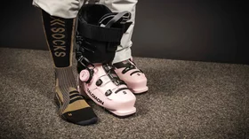 Ein Paar rosafarbene Salomon Skischuhe steht auf einem dunkelbraunen Teppich. Ein Bein, bekleidet mit einer hellgrauen Skihose und schwarzen Socken mit goldfarbener „XSOCKS“-Beschriftung, ist sichtbar. Der Skischuh ist geöffnet und zeigt das weiche Innenfutter. Das Bild strahlt eine Atmosphäre von Eleganz und Vorbereitung auf den Wintersport aus. Die Farbkombination aus Rosa, Schwarz und Gold wirkt modern und stilvoll. Der Fokus liegt auf den detailliert dargestellten Skischuhen und den hochwertigen Socken. Die Szene ist einfach, aber effektiv inszeniert und vermittelt ein Gefühl von Qualität und Funktionalität. © Sport Kessler GmbH