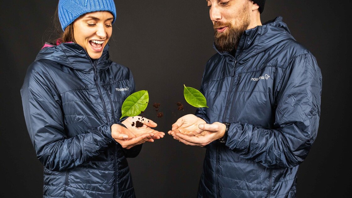 Ein Paar in dunkelblauen Outdoor-Jacken hält schwebende Kaffeebohnen und grüne Blätter in ihren Händen. © Sport Kessler GmbH