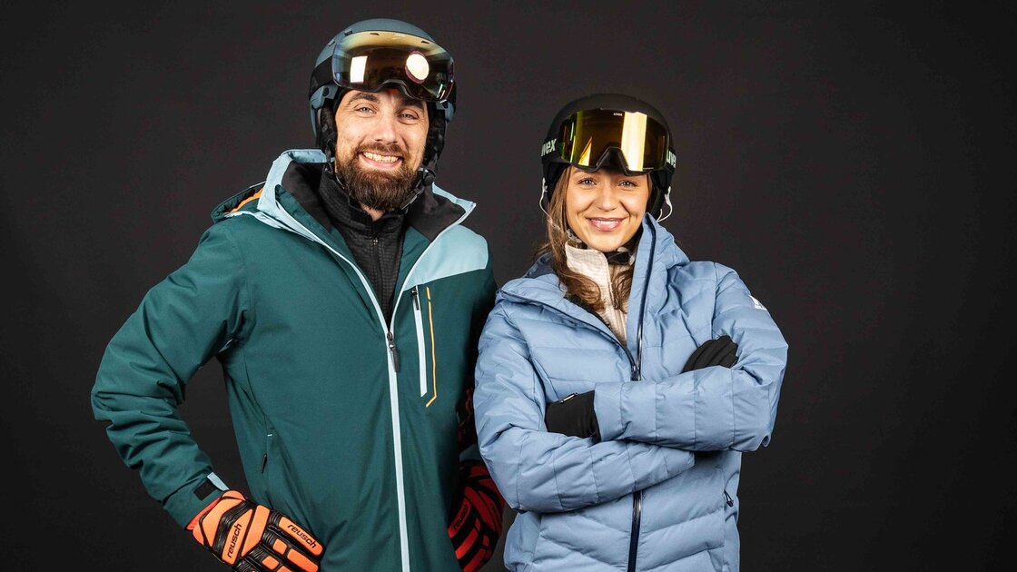 Ein glückliches Paar in kompletter, moderner Skiausrüstung, einschließlich Helm und Goggles, vor dunklem Hintergrund. © Sport Kessler GmbH