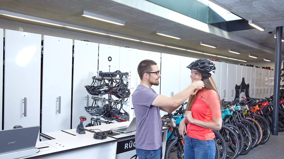 A male shop assistant helps a female customer adjust a bicycle helmet in a bright, modern bike shop. © Sport Kessler GmbH