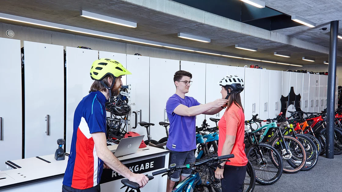 A staff member helps a customer try on a bike helmet in a bright, modern bike shop. Several e-bikes are lined up against a wall of lockers. © Sport Kessler GmbH