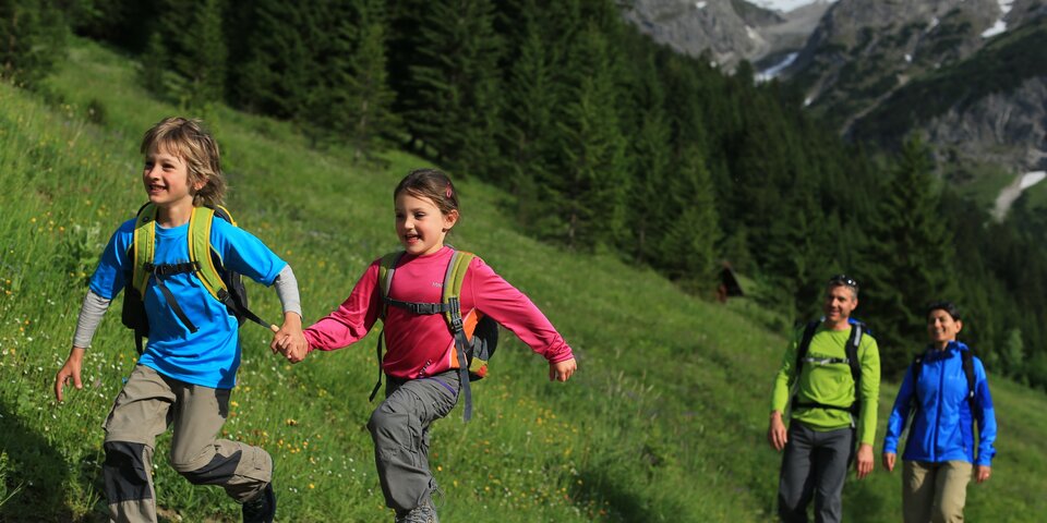 Zwei Kinder, Hand in Hand, rennen einen Bergweg entlang, während ihre Eltern ihnen folgen. &copy; Sport Kessler GmbH