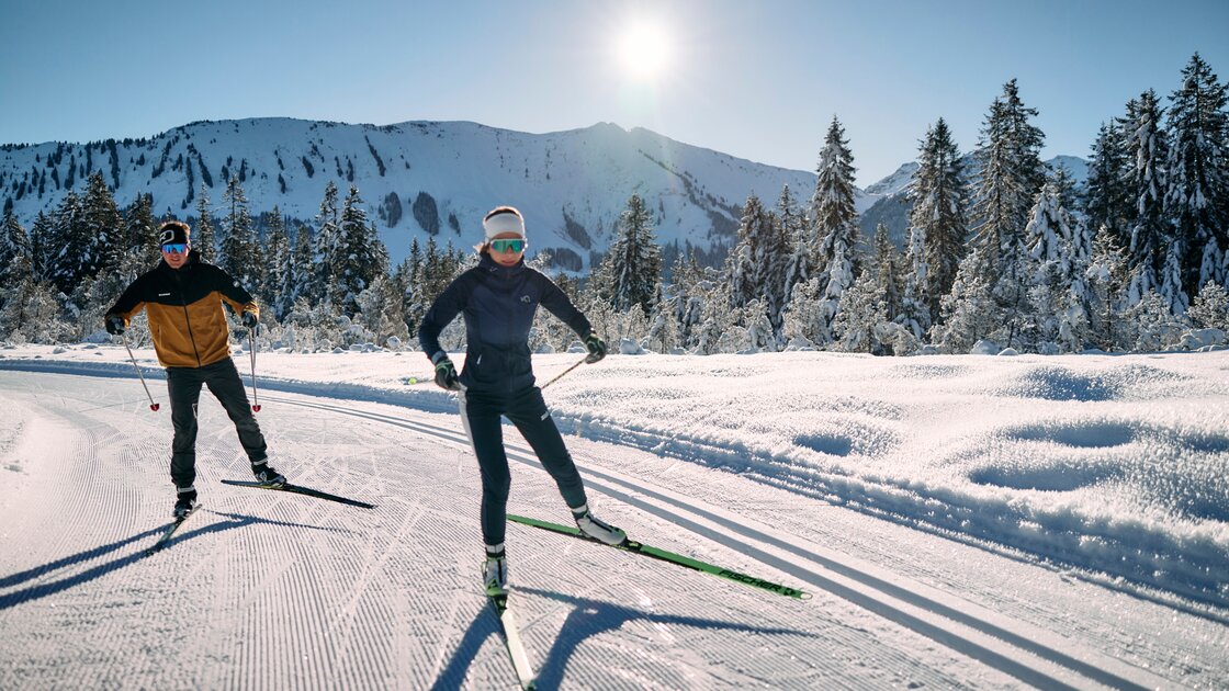 Zwei Langläufer trainieren auf einer präparierten Loipe vor einer schneebedeckten Bergkette. © Kleinwalsertal Tourismus eGen