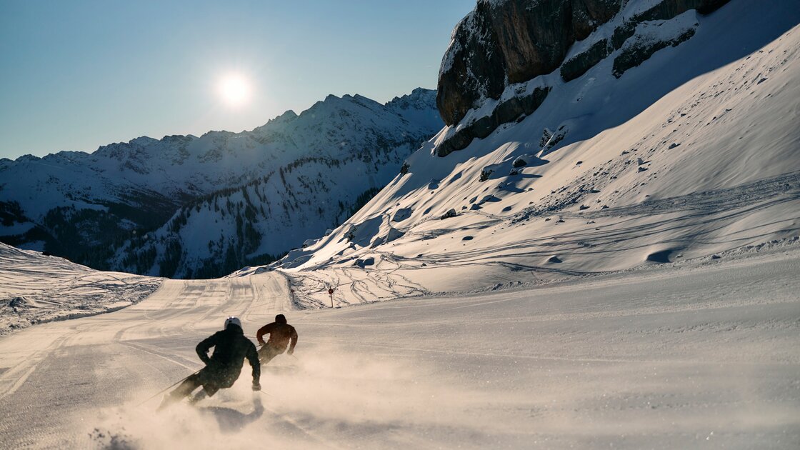Zwei Skifahrer fahren dynamisch eine sonnige Skipiste mit dramatischen Bergkulissen hinunter. © Kleinwalsertal Tourismus eGen