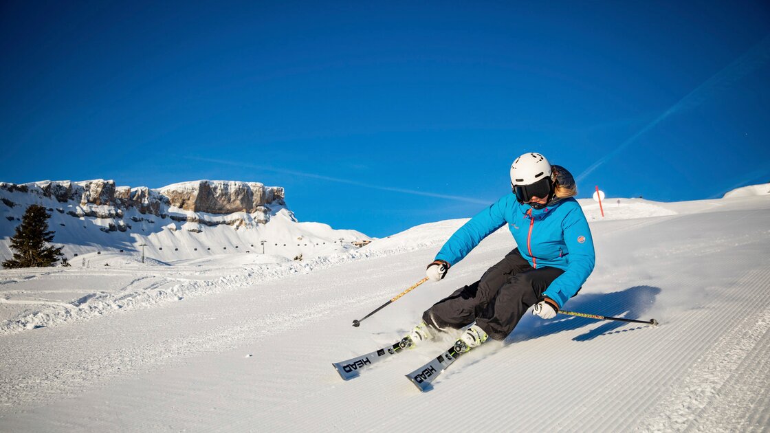 Skifahrerin in leuchtend blauer Jacke fährt eine präparierte Piste bei strahlend blauem Himmel hinunter. © Kleinwalsertal Tourismus eGen