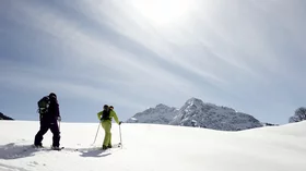 Zwei Skitourengeher wandern an einem sonnigen Wintertag durch eine verschneite Berglandschaft. Die Sonne scheint hell vom Himmel, der von zarten, streifigen Wolken durchzogen ist. Im Hintergrund erheben sich schneebedeckte Berge, deren Gipfel im gleißenden Licht erstrahlen. Die beiden Skitourengeher sind in funktioneller Winterkleidung gekleidet und scheinen auf ihrem Weg zu einem Gipfelziel konzentriert zu sein. Die Szene vermittelt ein Gefühl von Ruhe, Freiheit und der Schönheit der unberührten Natur. Die Weite der Landschaft und die klare Luft lassen die Stille der Berge spürbar werden. Das Bild ist geprägt von einem starken Kontrast zwischen dem hellen Schnee, dem blauen Himmel und den dunklen Silhouetten der Skifahrer, was die Klarheit und Weite des Motivs unterstreicht. © Sport Kessler GmbH