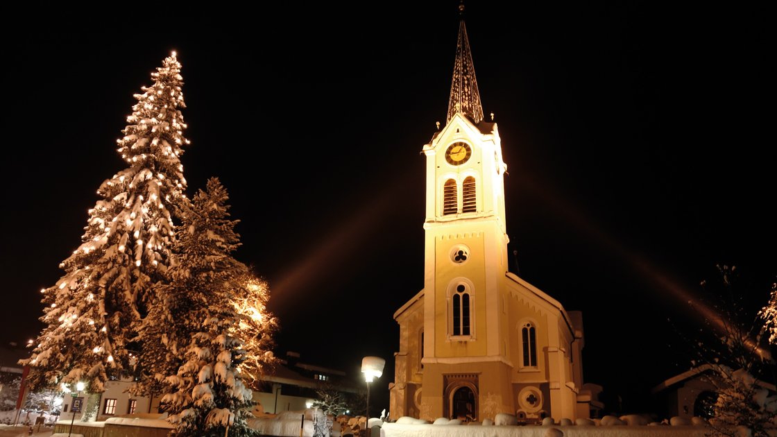 Nighttime view of a snow-covered church and Christmas tree. &copy; Kleinwalsertal Tourismus eGen