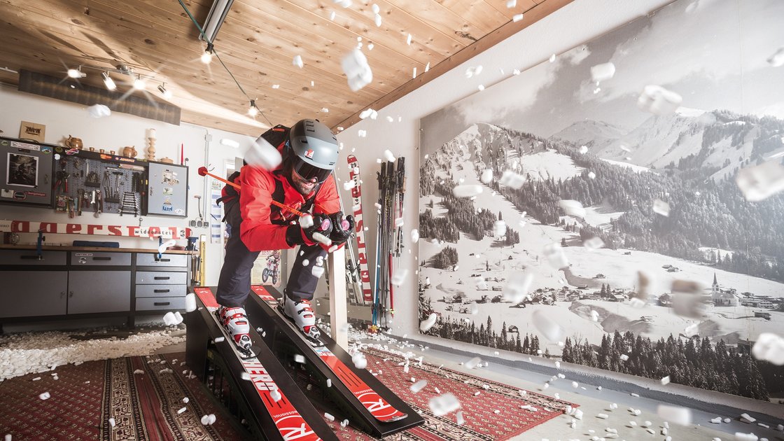 Skifahrer in einer Garage, umgeben von Styroporkügelchen, die in der Luft schweben. Er steht auf Skiern auf einer kleinen Rampe, die in einem Raum mit Werkbank und Skiausrüstung aufgestellt ist. An der Wand hängt ein großes Schwarz-Weiß-Foto einer verschneiten Berglandschaft. Das Bild vermittelt den Eindruck von Bewegung und Dynamik, trotz des ungewöhnlichen Umfelds. Der Skifahrer scheint sich inmitten eines Schneesturms zu befinden, obwohl sich der Schnee aus Styroporkügelchen zusammensetzt. Die Szene ist surreal und humorvoll, zeigt aber gleichzeitig die Sehnsucht nach Schnee und Wintersport. Das Bild könnte als Kommentar zur Sehnsucht nach dem Winter, der Natur und dem Wintersport interpretiert werden, obwohl man sich in einer ganz anderen Umgebung befindet. © Sport Kessler GmbH
