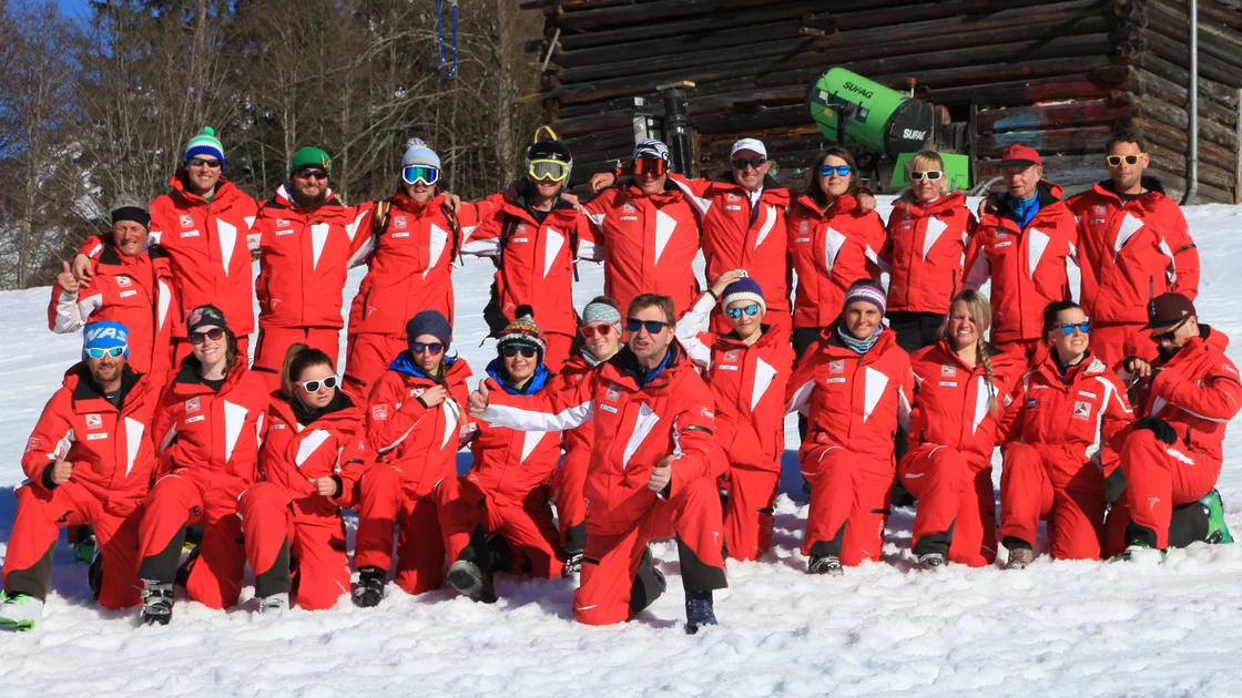 Eine Skilehrergruppe in roten Uniformen posiert im Schnee vor einem Holzhaus. © Skischule Seite Egg